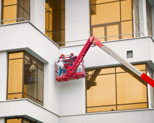 Construction Workers Working At Height In Lifting Bucket. Plasterer Worker In Hardhat Paints Facade Of The Building At Height In The Cradle. Finishing Facade Work