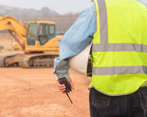 An Engineer Or Worker Wearing A Life Jacket Holds A Radio Transmitter And A White Helmet In His Hands. For Safe Working On Excavator Background